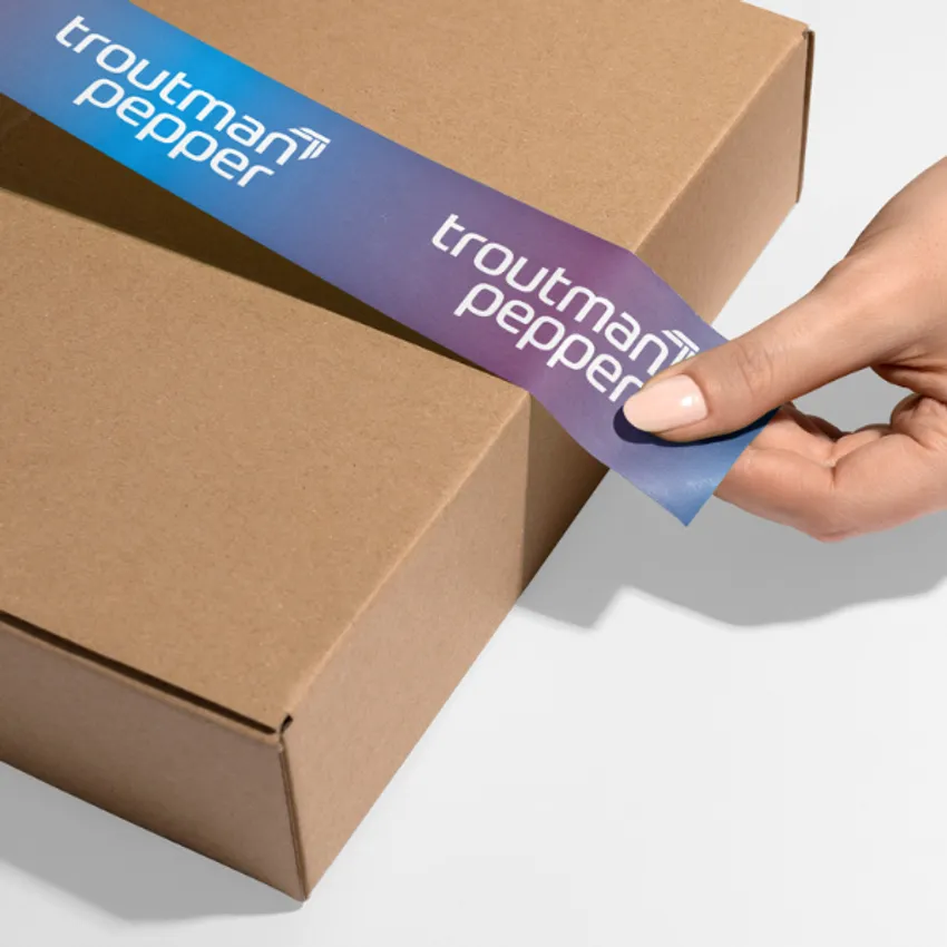 Hands applying custom branded shipping tape for Troutman Pepper onto a shipping box on a white background